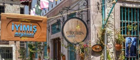 A cobblestone street with a restaurant named "İYIMİS meyhanesi" and another named "Orchis". There are potted plants and a chalkboard sign with menu items.