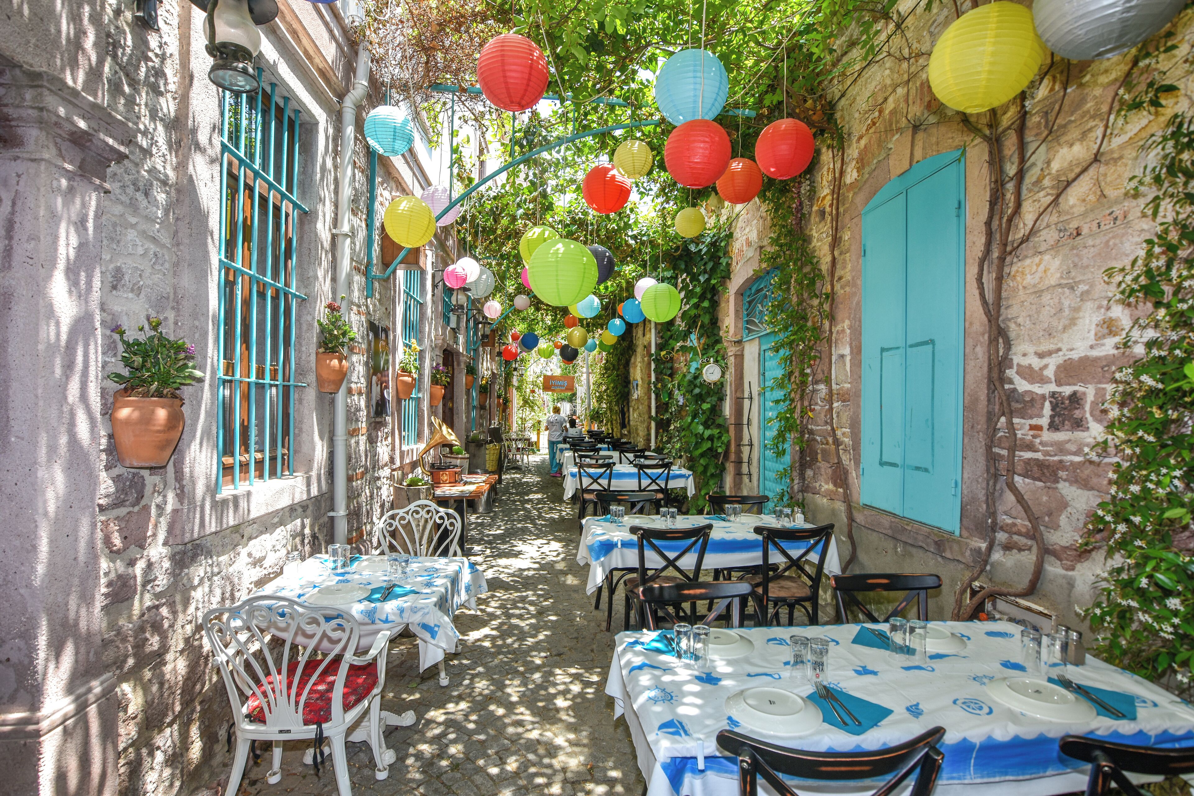 A narrow alley with tables and chairs set up for dining, flanked by buildings with blue shutters and hanging lanterns.