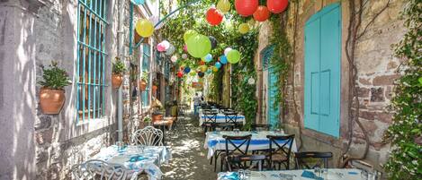 A narrow alley with tables and chairs set up for dining, flanked by buildings with blue shutters and hanging lanterns.