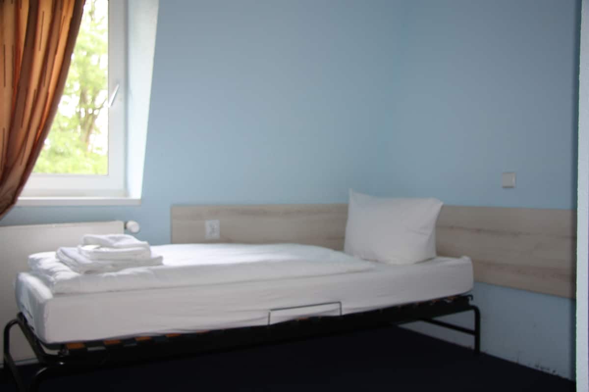 A single bed with white bedding in a room with light blue walls and a window with curtains.