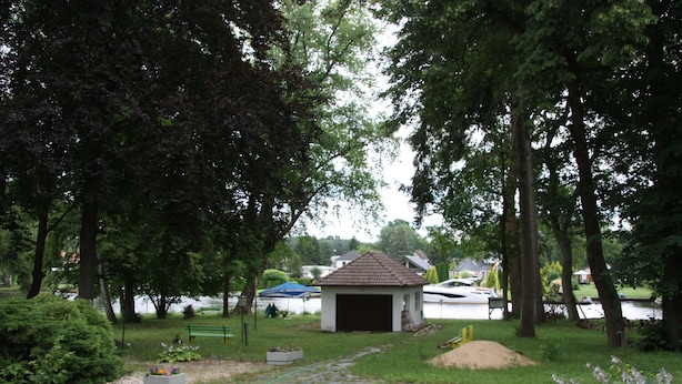 A small building with a tiled roof, surrounded by trees and a grassy area with a pathway leading to it. - Hotel am Fluss (Heidesee)