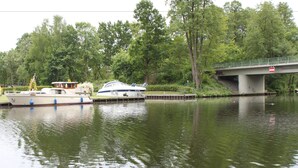 A calm river with boats docked on the left, a bridge in the background, and lush greenery surrounding the area. - Hotel am Fluss (Heidesee)