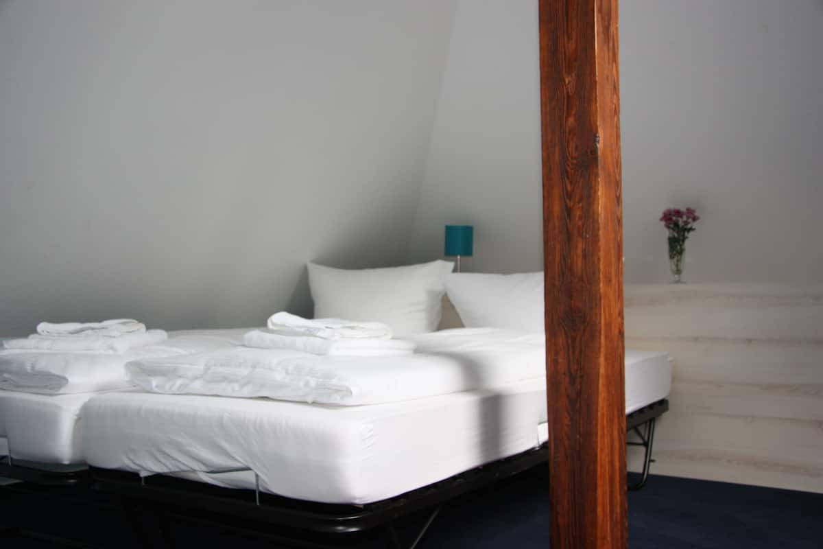 A neatly made bed with white linens and pillows, a wooden bed frame, a teal nightstand with a lamp, and a small vase with flowers on the floor.
