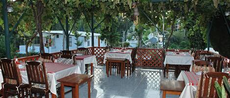 Un área de comedor al aire libre con mesas y sillas de madera, cubierta por una pérgola con enredaderas de uva.
