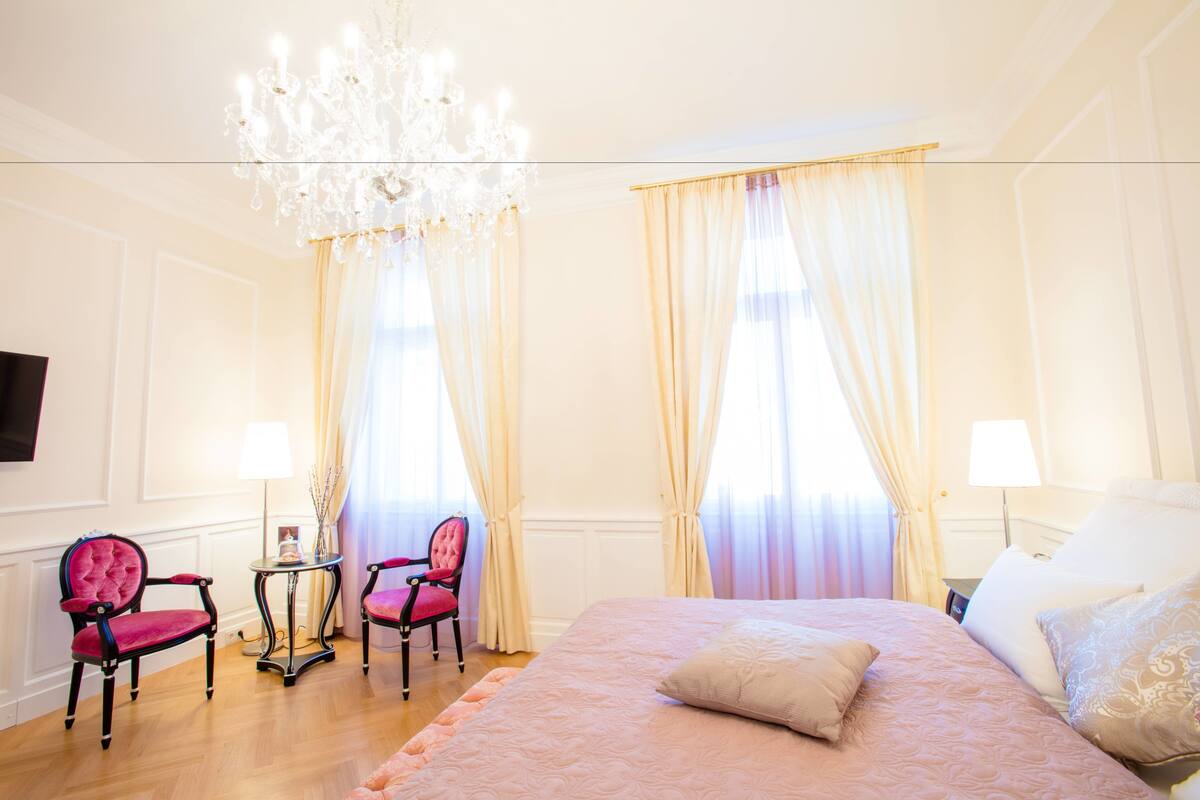 Ein Hotelzimmer mit einem großen Bett, zwei Sesseln, einem kleinen Tisch, einer Chandelüster und Vorhängen an den Fenstern.