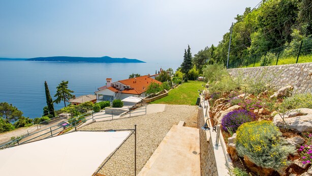 A coastal property with a stone retaining wall, a garden with various plants, and a view of the sea and distant hills. - Apartments Gea (Moscenicka Draga)