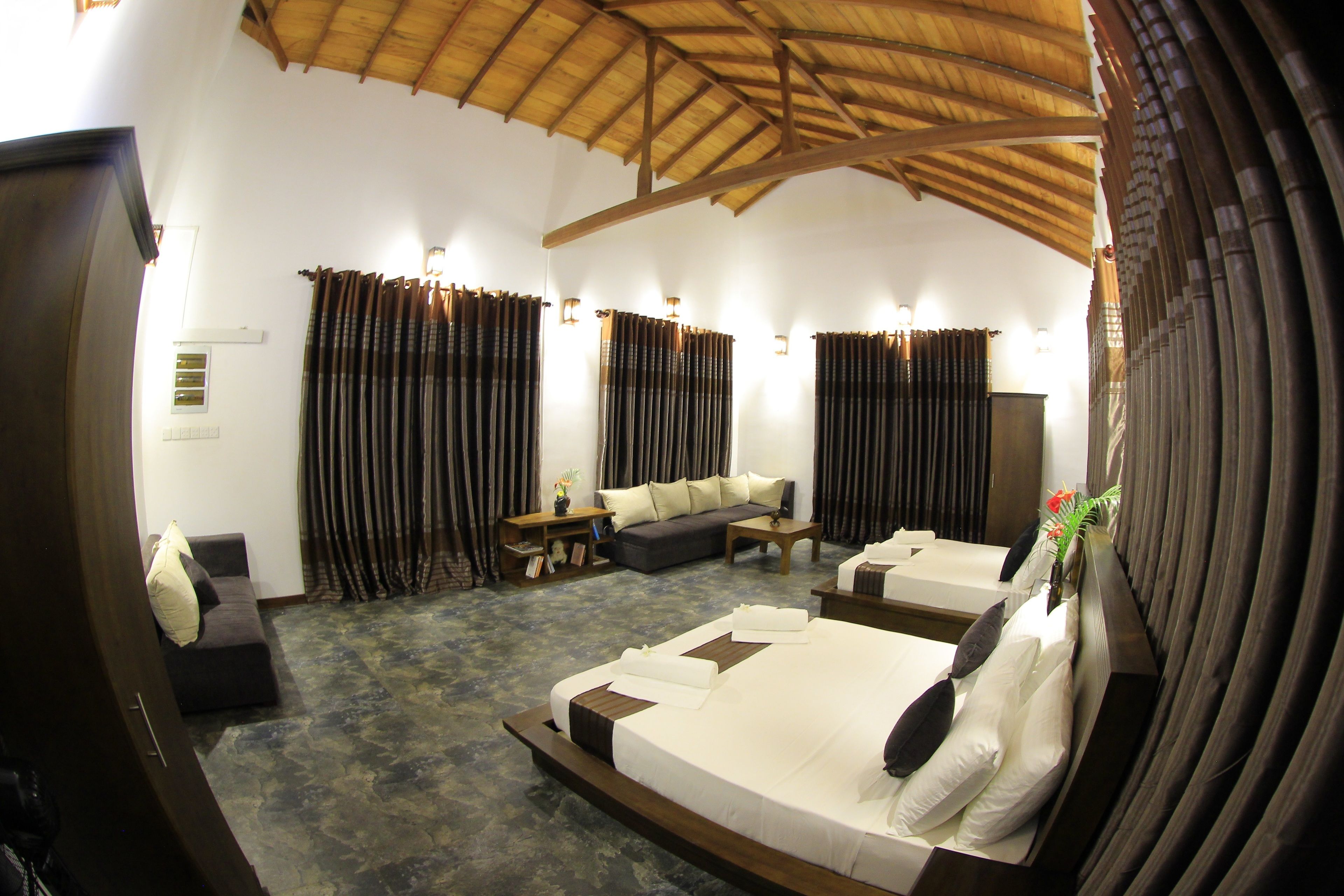 A spacious room with a large bed, a sofa, and a small table. There are curtains on the windows and a ceiling with wooden beams.