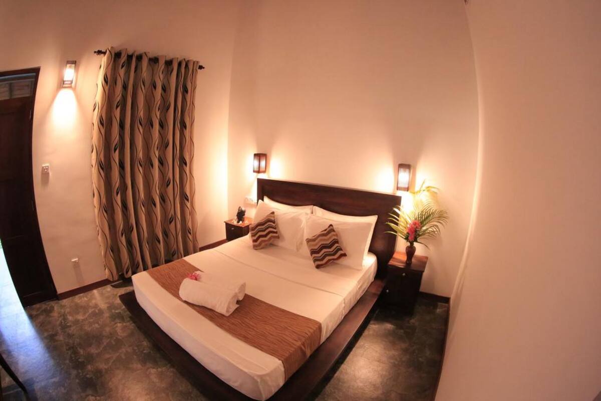 A hotel room with a large bed, two bedside lamps, a nightstand with a lamp, a curtain, and a potted plant.
