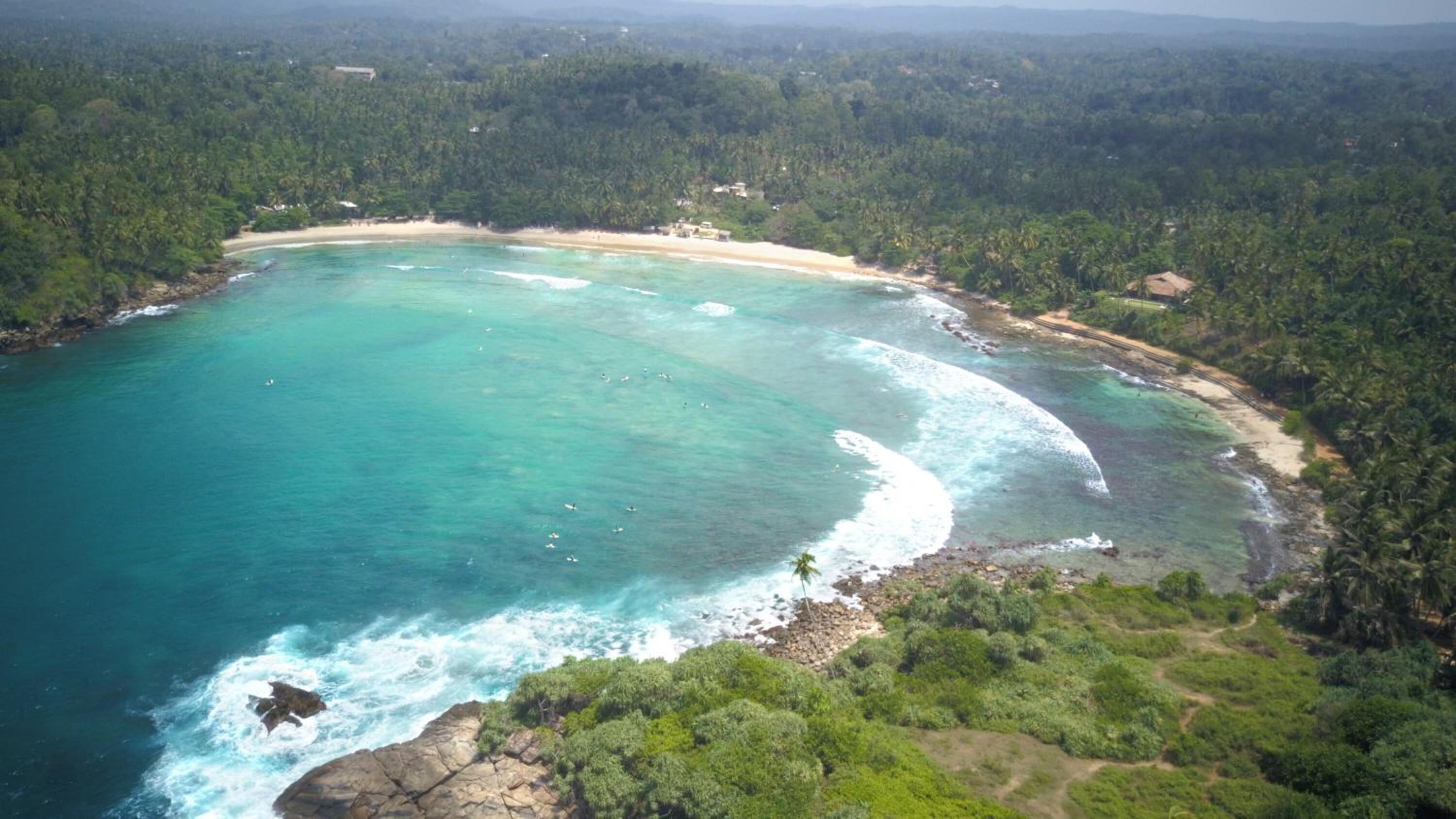 An aerial view of a coastal area with a beach, dense forest, and clear turquoise waters.