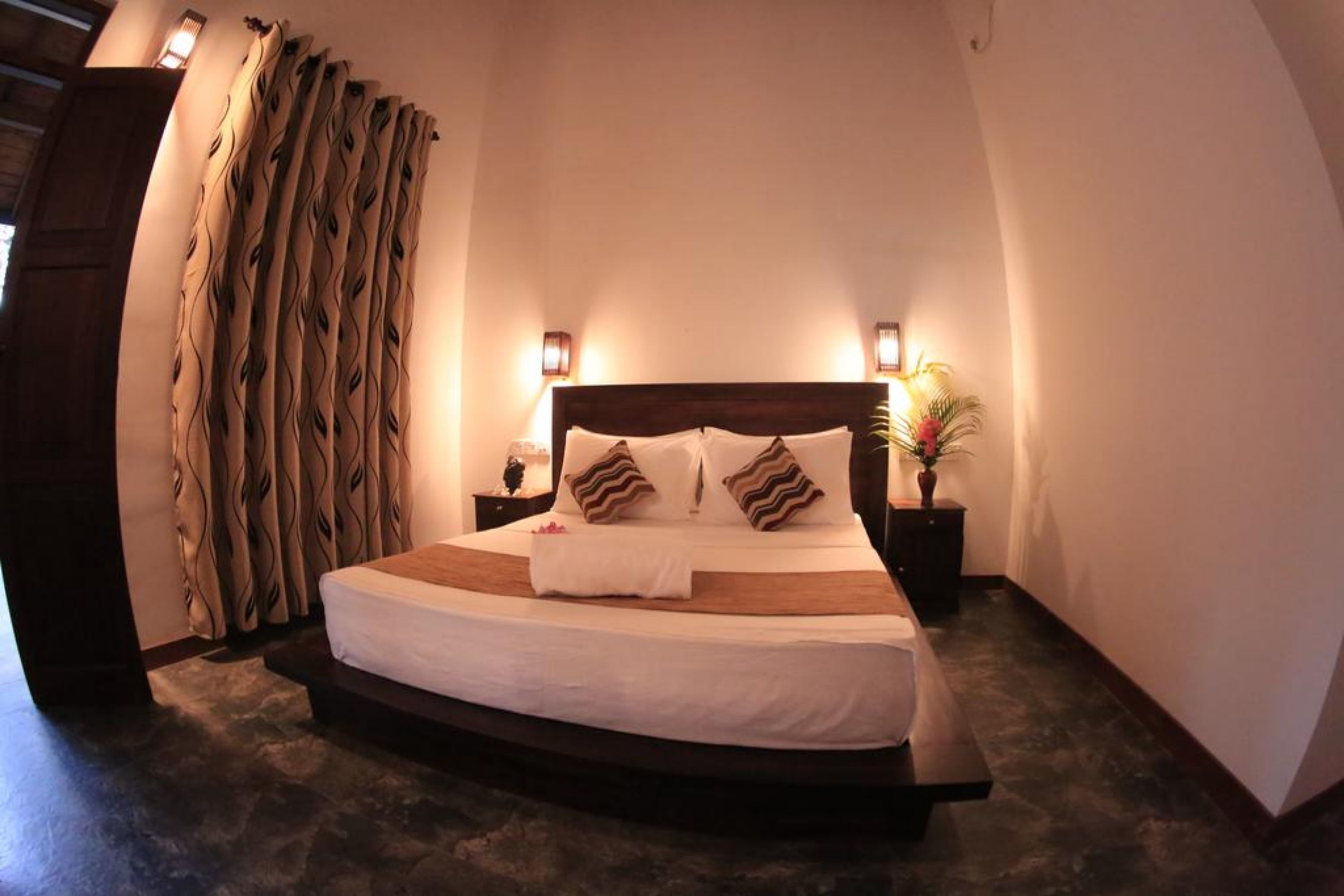 Quarto com teto arredondado, uma cama com cabeceira de madeira, duas mesas de cabeceira com abajures e uma janela grande com cortinas.