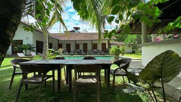 A poolside area with a table and chairs, surrounded by lush greenery and a building with a tiled roof.
