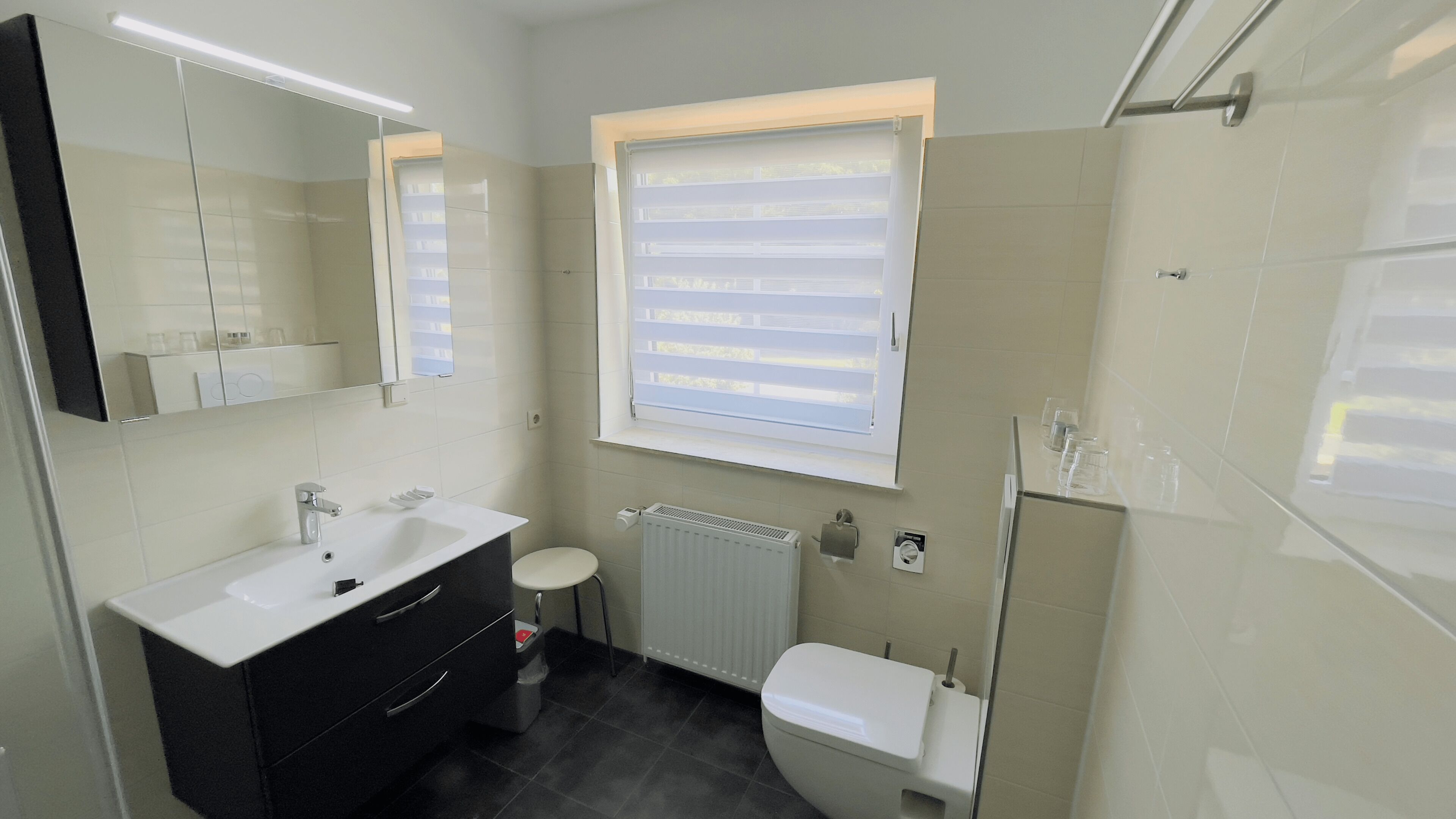 Un baño moderno con un lavamanos blanco, tocador negro y un inodoro. Hay una ventana con persianas, un radiador y un espejo sobre el lavamanos.