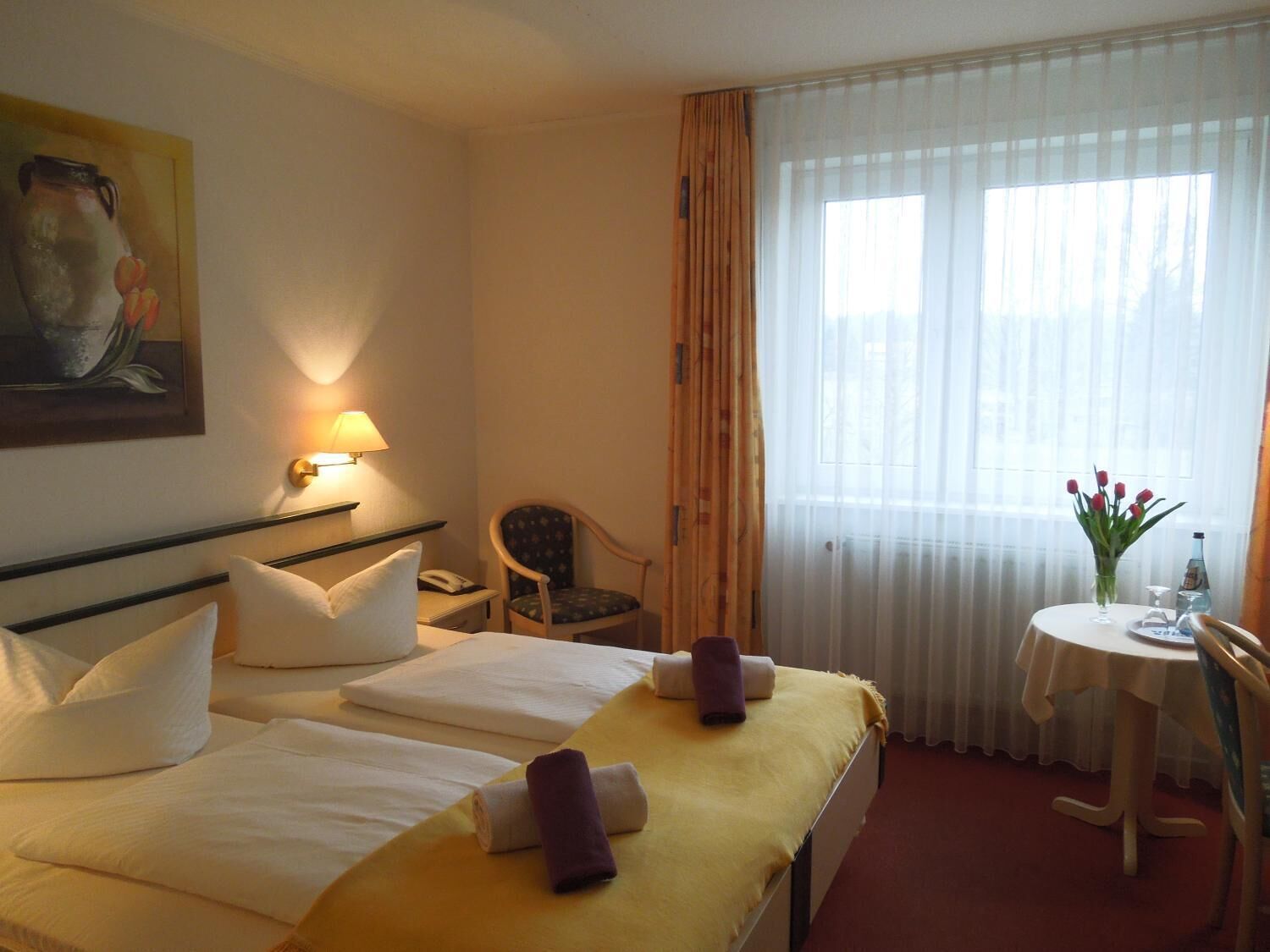 Habitación de hotel con dos camas, un escritorio, una silla, una lámpara, un cuadro y una ventana con cortinas.