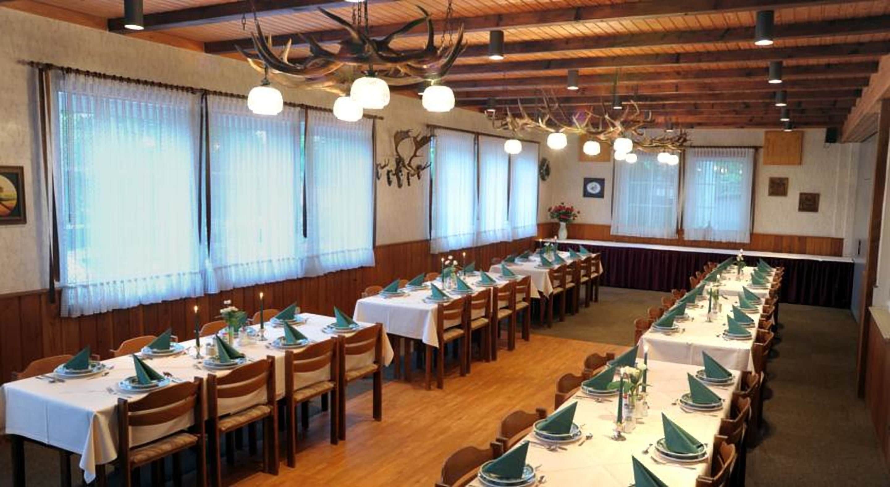A dining hall with long tables set for a meal, wooden ceiling beams, and large windows with sheer curtains.