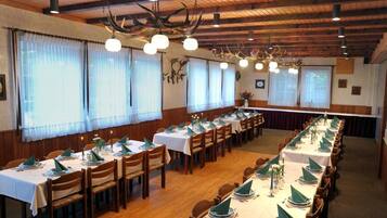 A dining hall with long tables set for a meal, wooden ceiling beams, and large windows with sheer curtains.