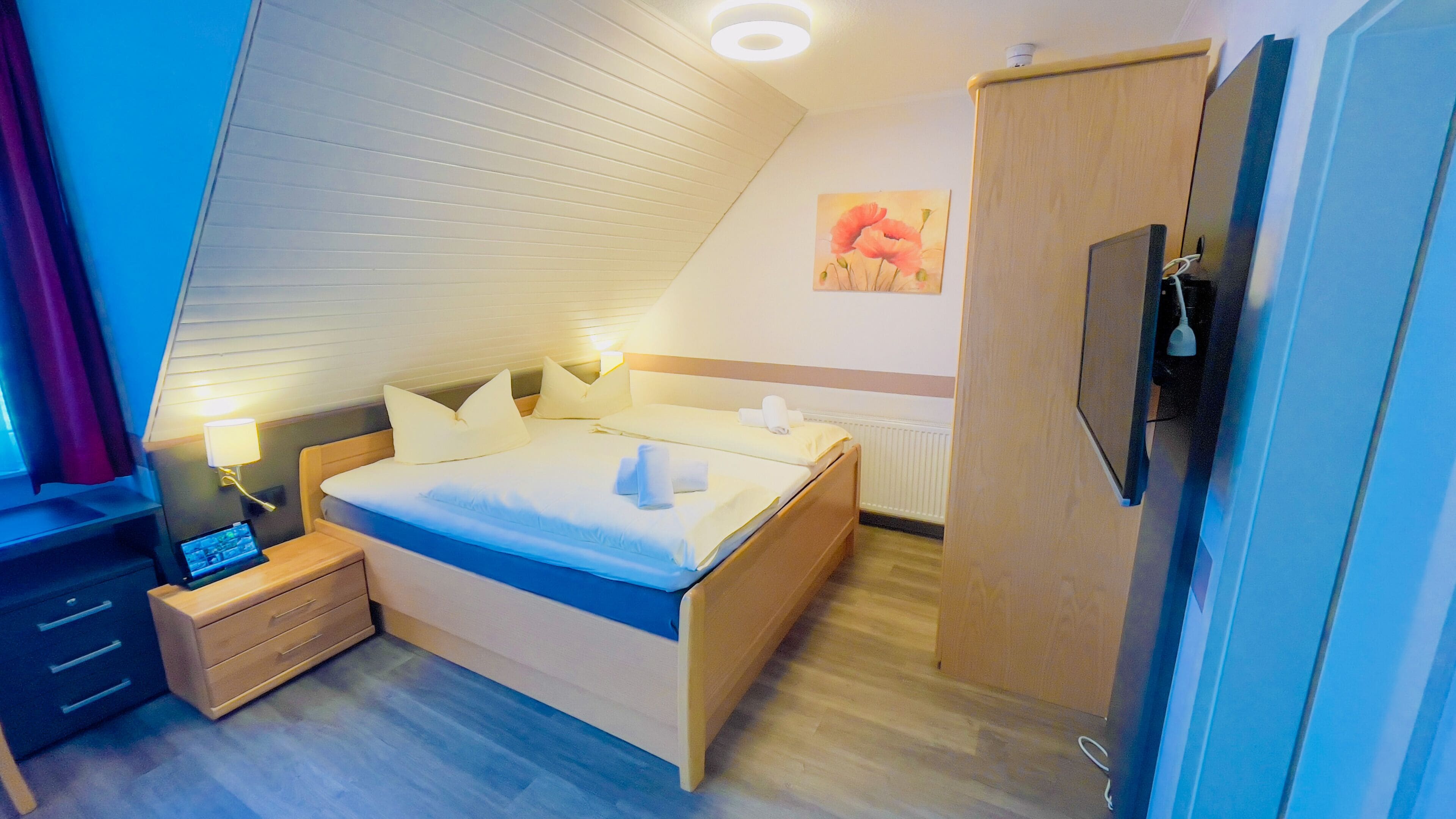 A modern hotel room with a sloped ceiling, a bed with white linens, a bedside table with a lamp, a TV, and a wooden wardrobe.