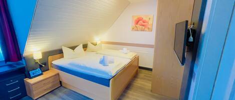 Una habitación de hotel moderna con techo inclinado, una cama con ropa blanca, una mesita de noche con lámpara, un televisor y un armario de madera.