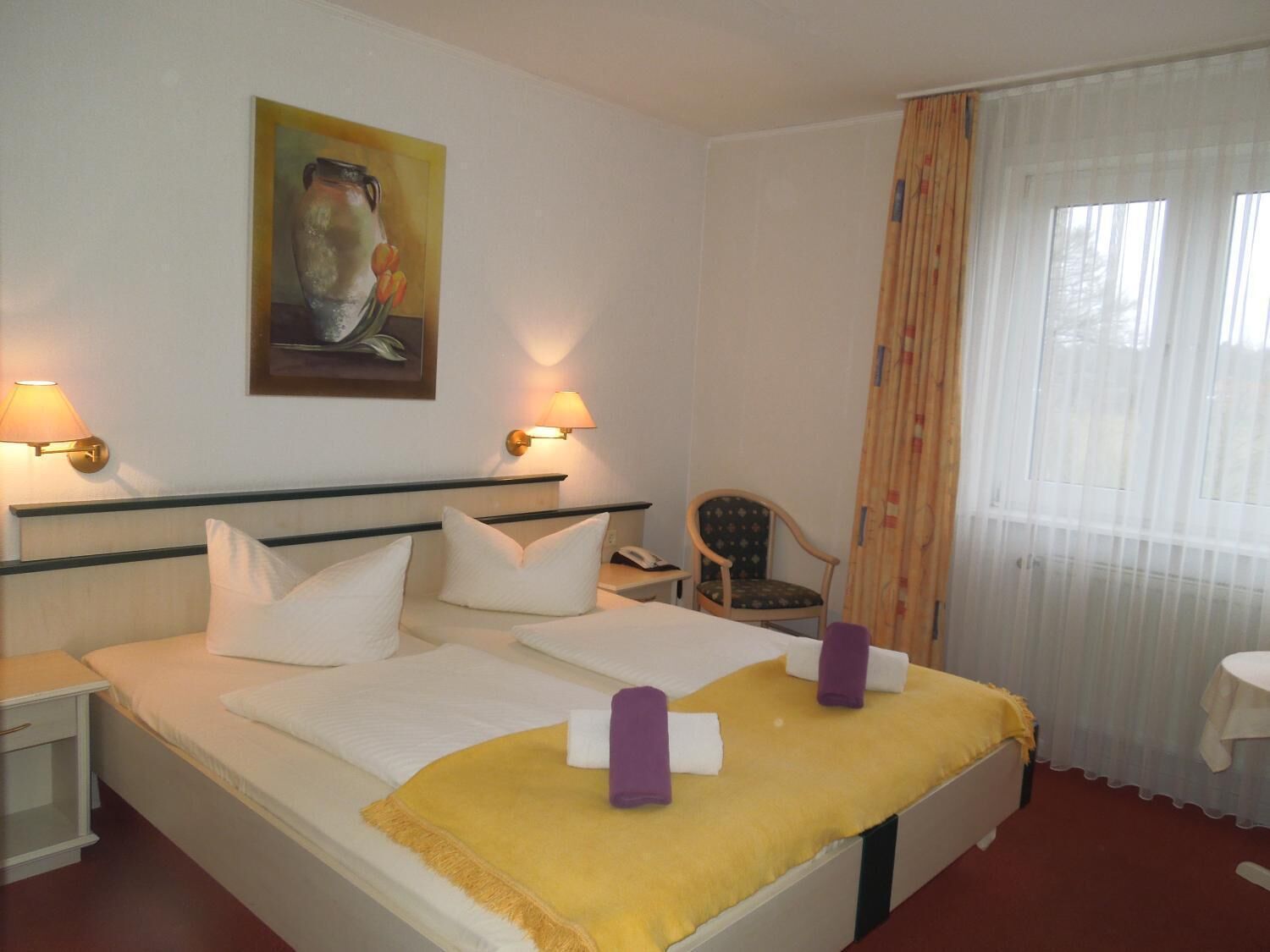 Una habitación de hotel con una cama, dos lámparas de noche, una silla, una mesita y un cuadro en la pared.