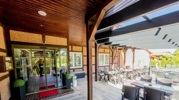 An outdoor patio area with tables and chairs, a glass entrance, and a wooden ceiling. - Hotel Schnehagen (Bad Fallingbostel)