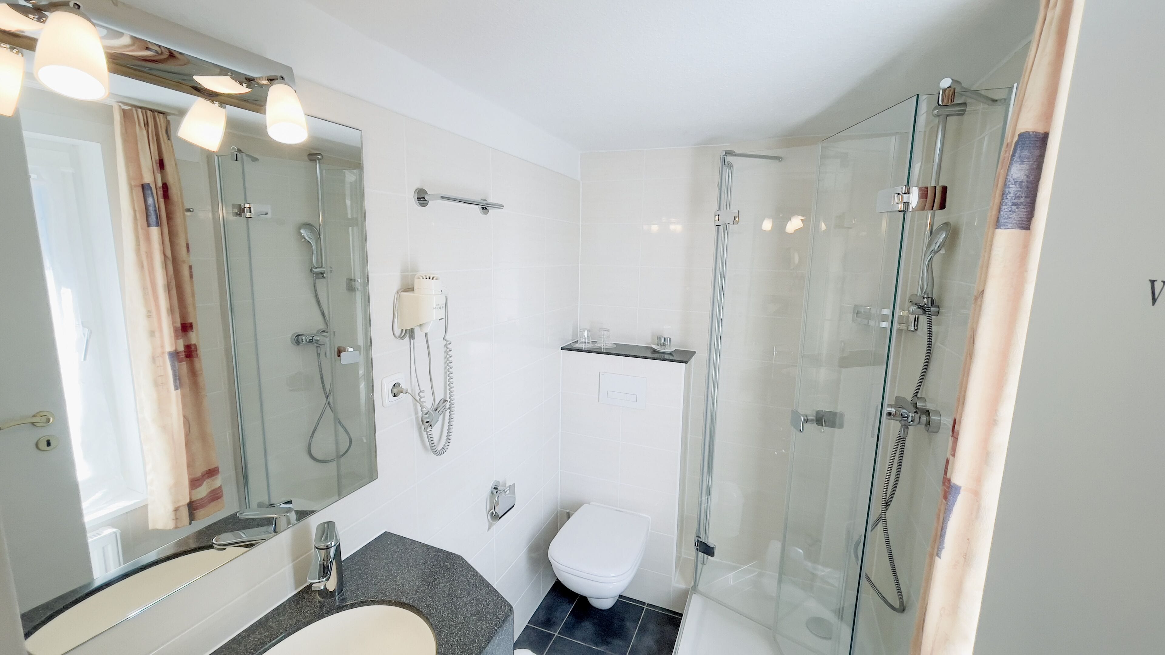 Un baño moderno con ducha de vidrio, inodoro, lavabo con espejo y secador de pelo fijado en la pared.