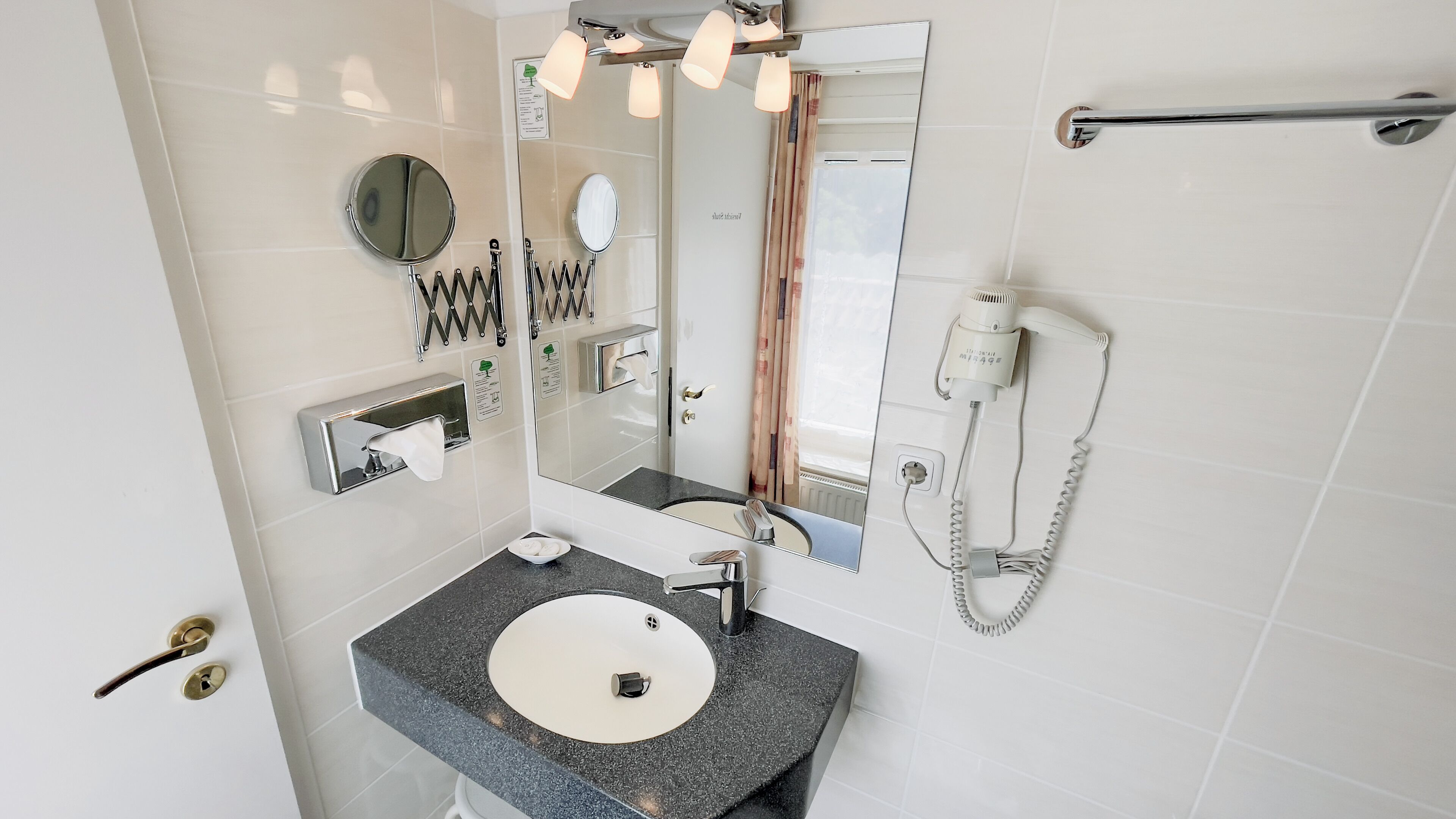 Een moderne badkamer met een wastafel met granieten blad, een spiegel, een handdoekdispenser en een haardroger.