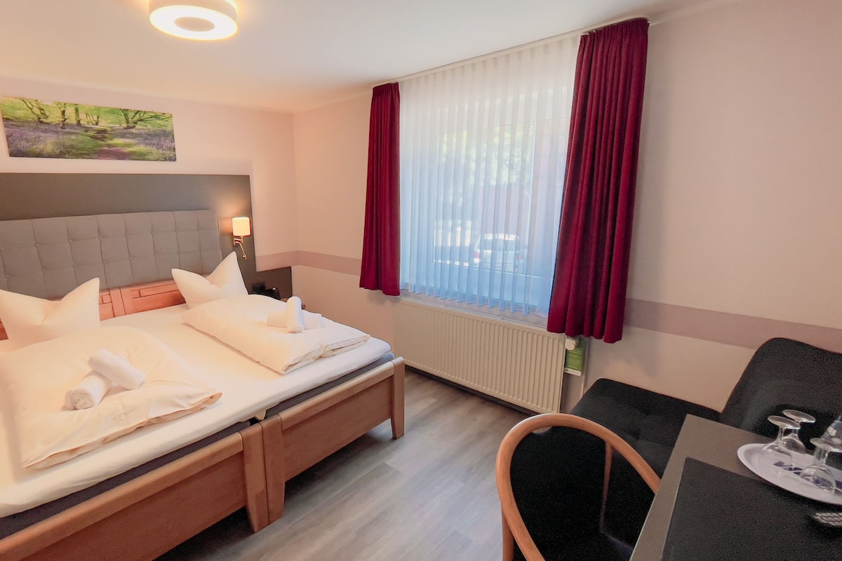 Quarto de hotel com cama, abajures, um quadro na parede, janela com cortinas e uma mesinha com copos.