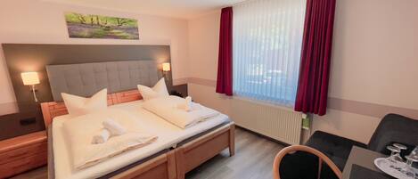 A hotel room with a bed, bedside lamps, a painting on the wall, a window with curtains, and a small table with glasses.