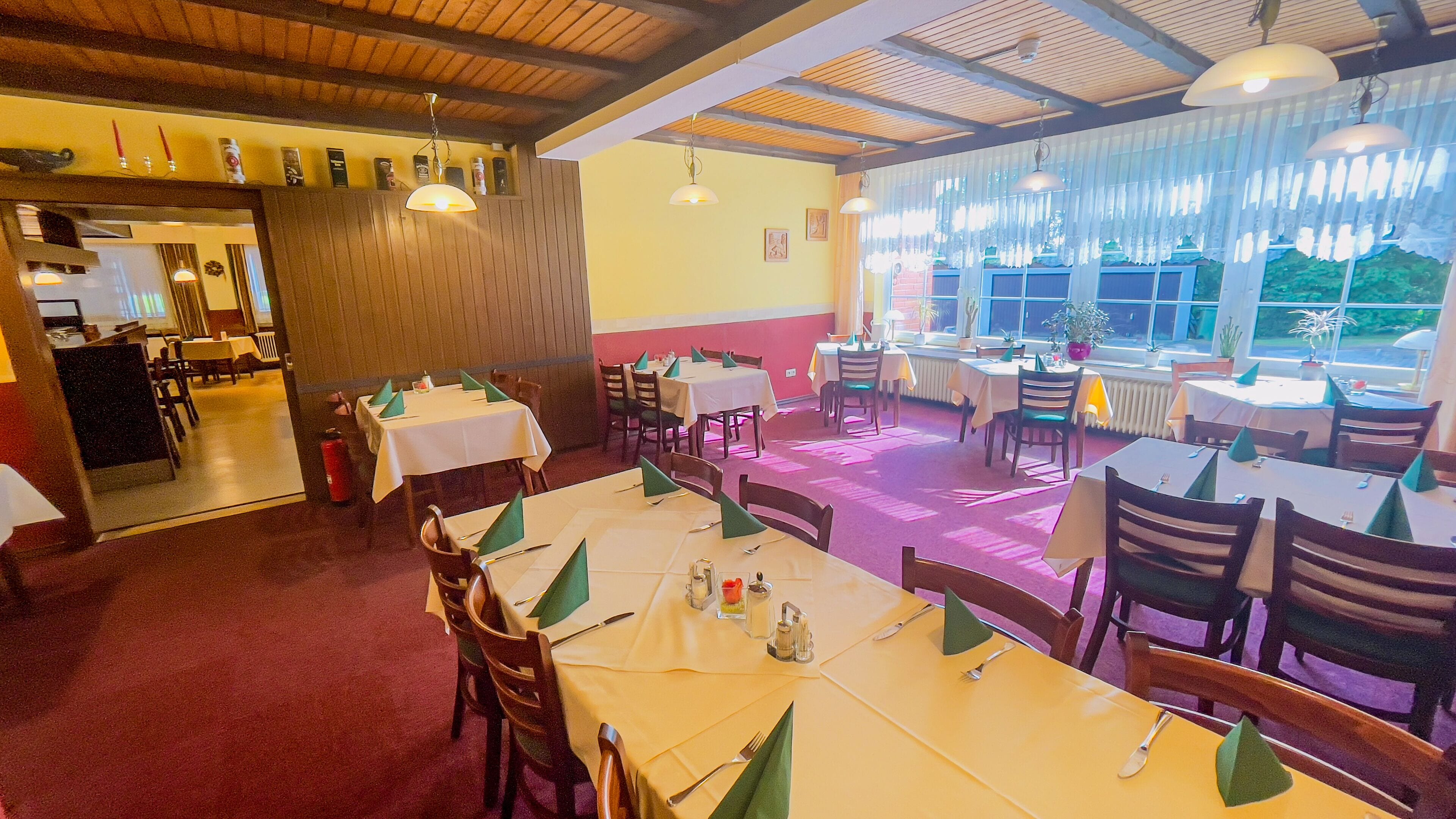 Ein Restaurant mit mehreren Esstischen, Holzbalkendecke und großen Fenstern mit Vorhängen.