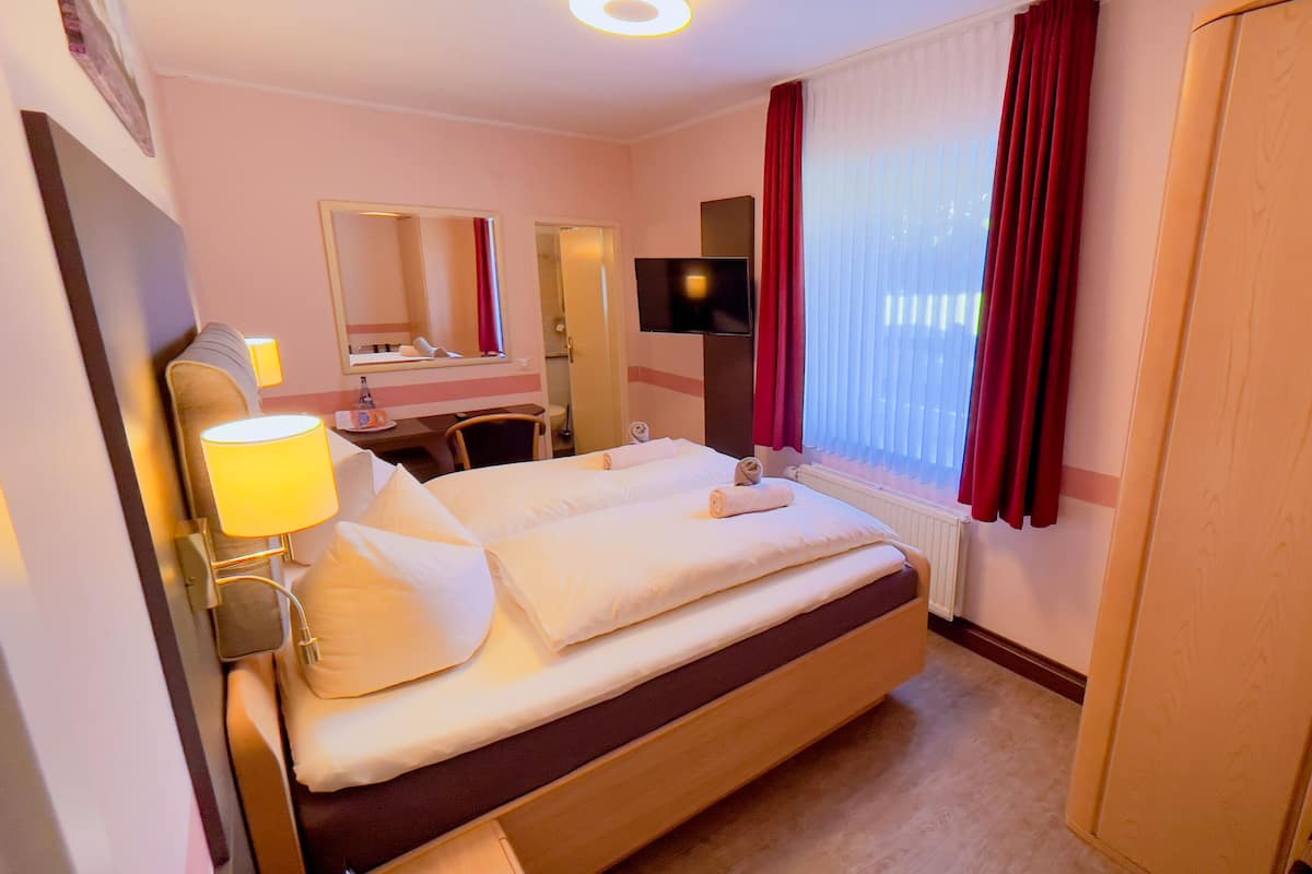 Quarto de hotel com cama, mesa de cabeceira, luminária, espelho, televisão e janela com cortinas.