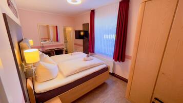A hotel room with a bed, bedside table, lamp, mirror, TV, and a window with curtains.