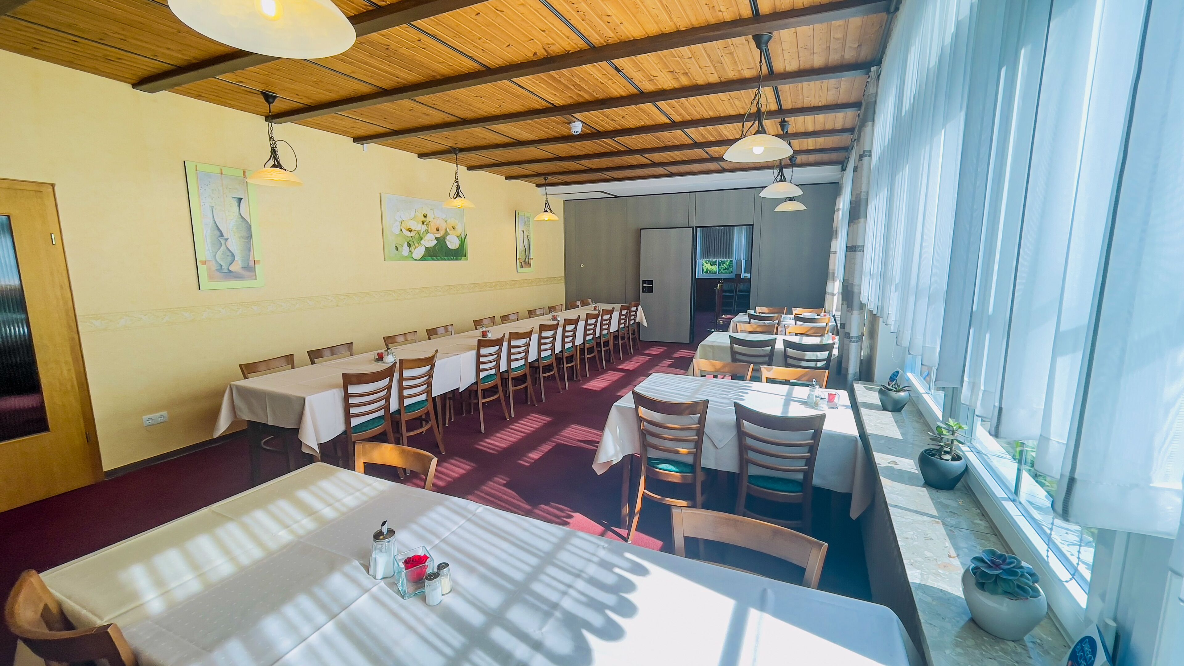 A dining area with multiple tables and chairs, wooden ceiling beams, and large windows allowing natural light.