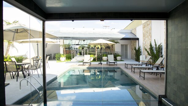 A modern outdoor pool area with glass walls, lounge chairs, and umbrellas. - Hotel Florença (Americana)