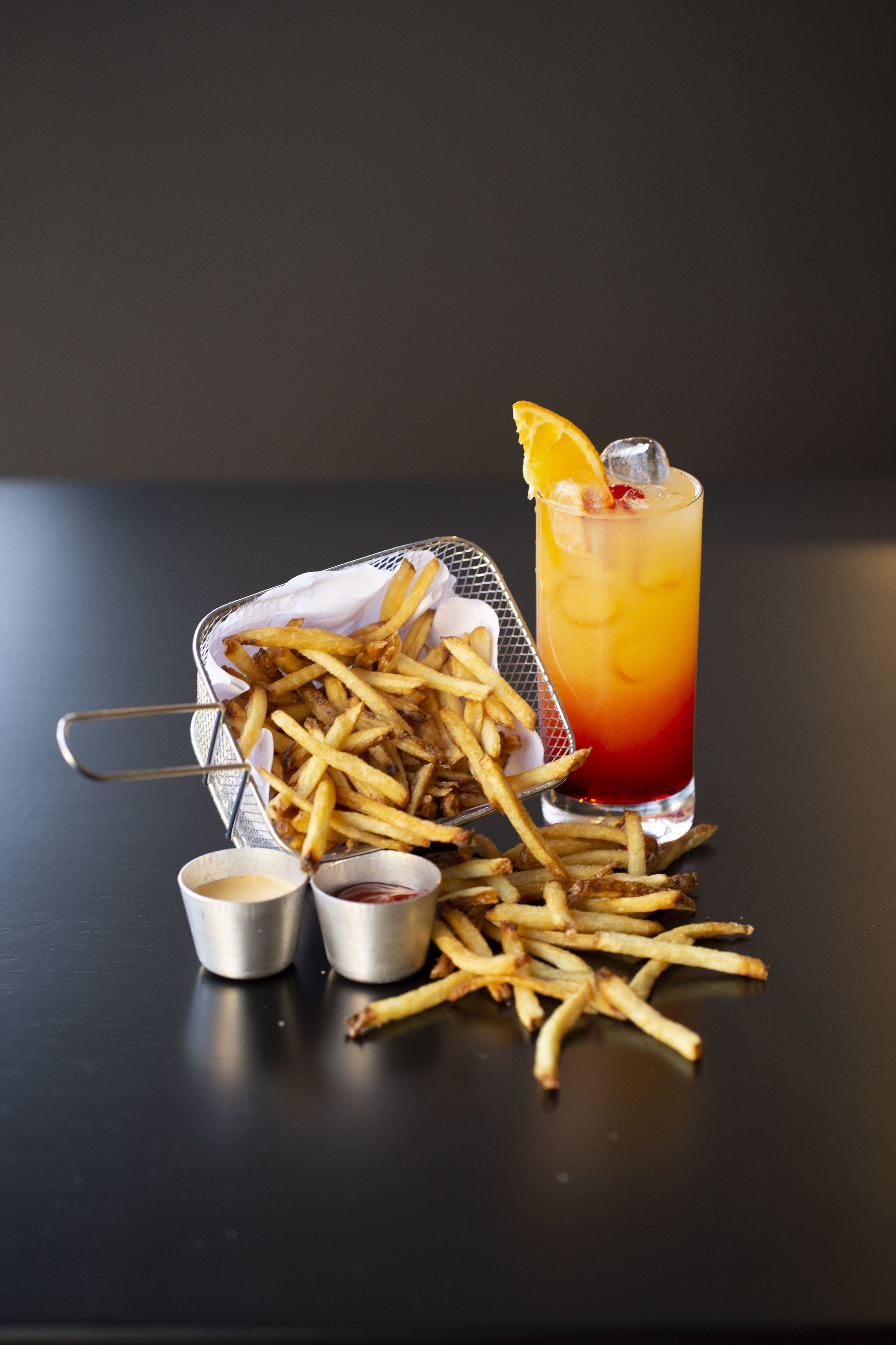 A basket of French fries, a tall glass of iced drink with a slice of orange, and two small containers of sauces on a dark surface.