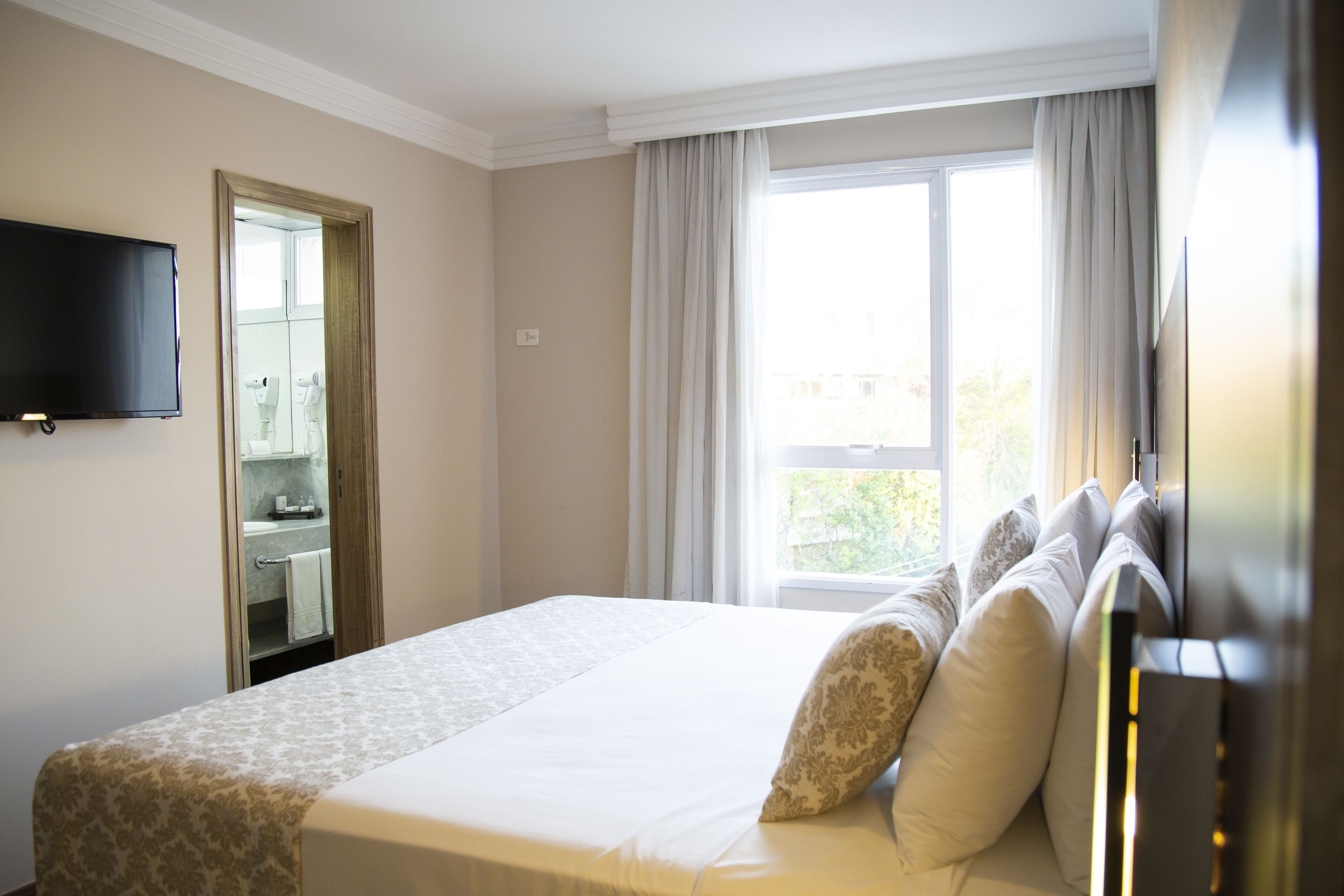 Una habitación de hotel con una cama, un televisor montado en la pared, una ventana con cortinas y un baño visible a través de una puerta abierta.