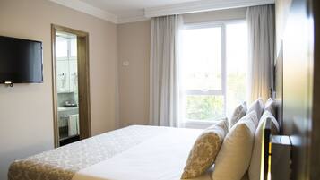 Una habitación de hotel con una cama, un televisor montado en la pared, una ventana con cortinas y un baño visible a través de una puerta abierta.