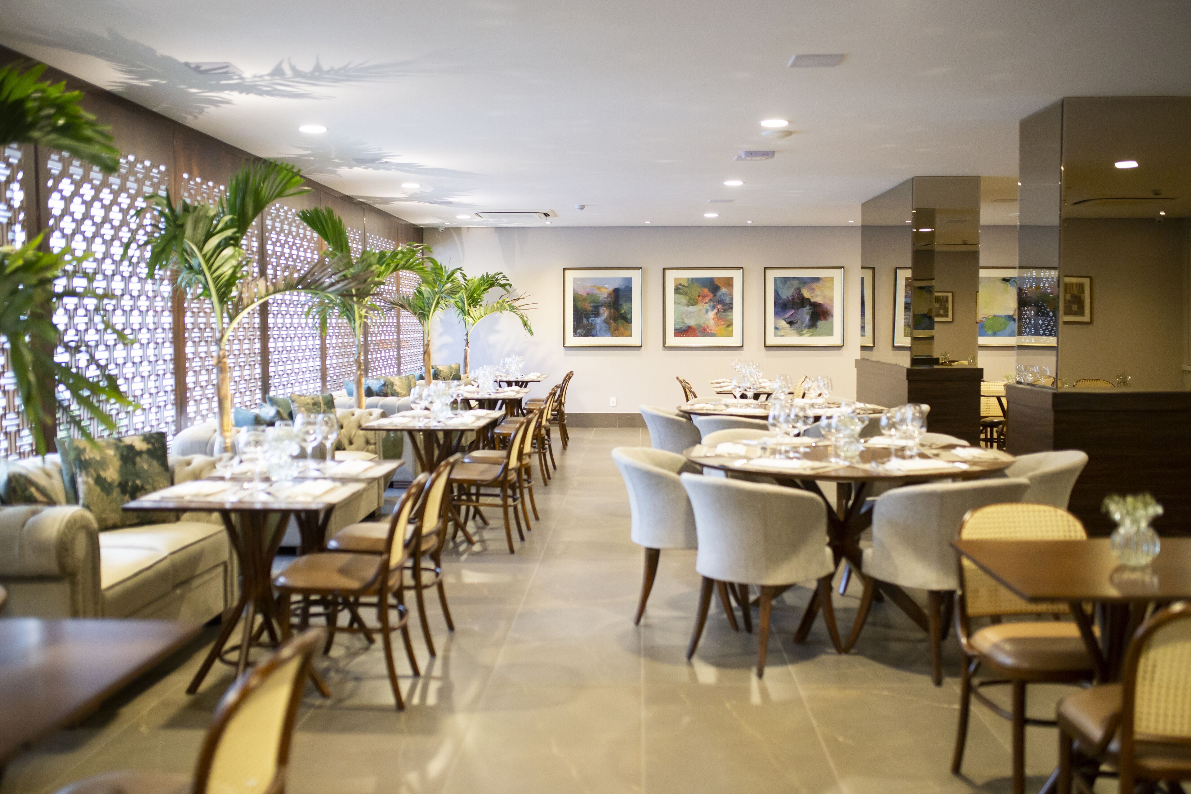Comedor con varias mesas dispuestas para recibir a los invitados, con un diseño moderno, sillas de colores claros y una pared decorativa con obras de arte enmarcadas.