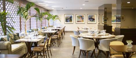 Comedor con varias mesas dispuestas para recibir a los invitados, con un diseño moderno, sillas de colores claros y una pared decorativa con obras de arte enmarcadas.