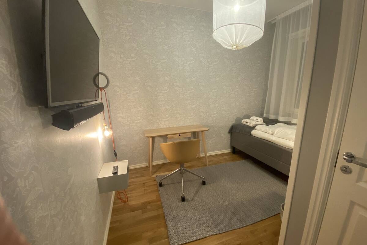 Une chambre à coucher comprenant un lit, un bureau, une chaise, un téléviseur fixé au mur et un luminaire suspendu.