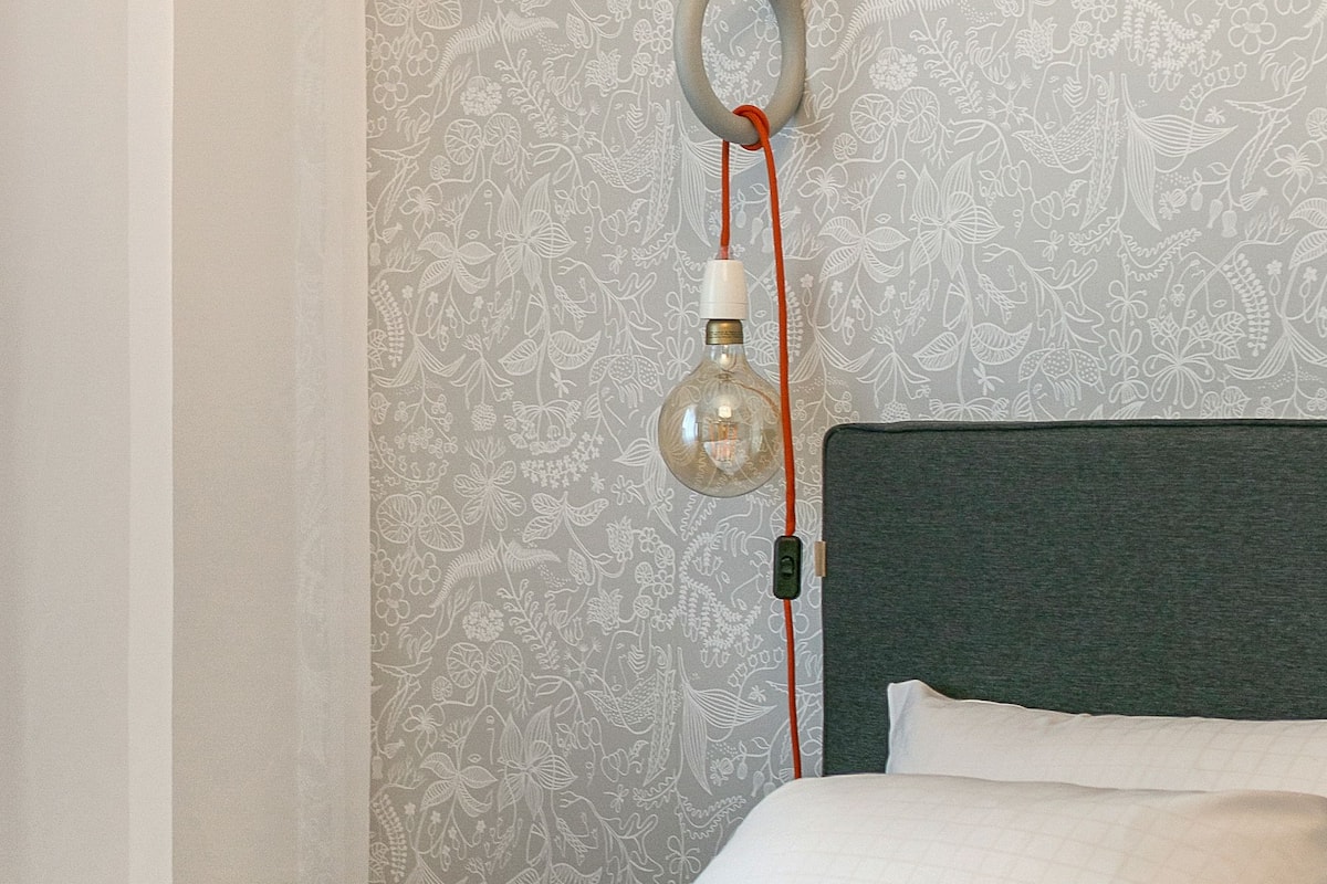 Un lit bien fait, avec du linge de lit blanc, une tête de lit grise et un mur recouvert d’un papier peint à motifs floraux.