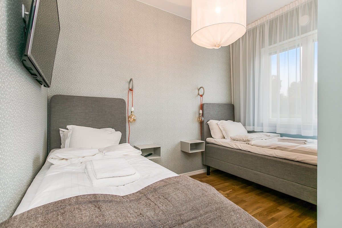 Une chambre d’hôtel moderne avec un grand lit, un petit canapé, un téléviseur fixé au mur et une fenêtre ornée de rideaux légers.