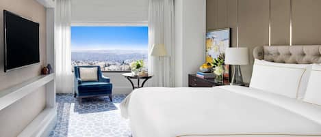 A hotel room with a large bed, a TV, a chair, a small table, and a view of the city.