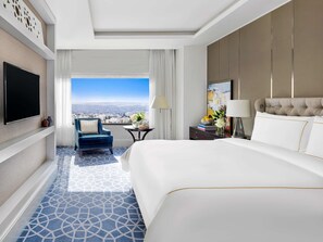 A hotel room with a large bed, a TV, a chair, a small table, and a view of the city.