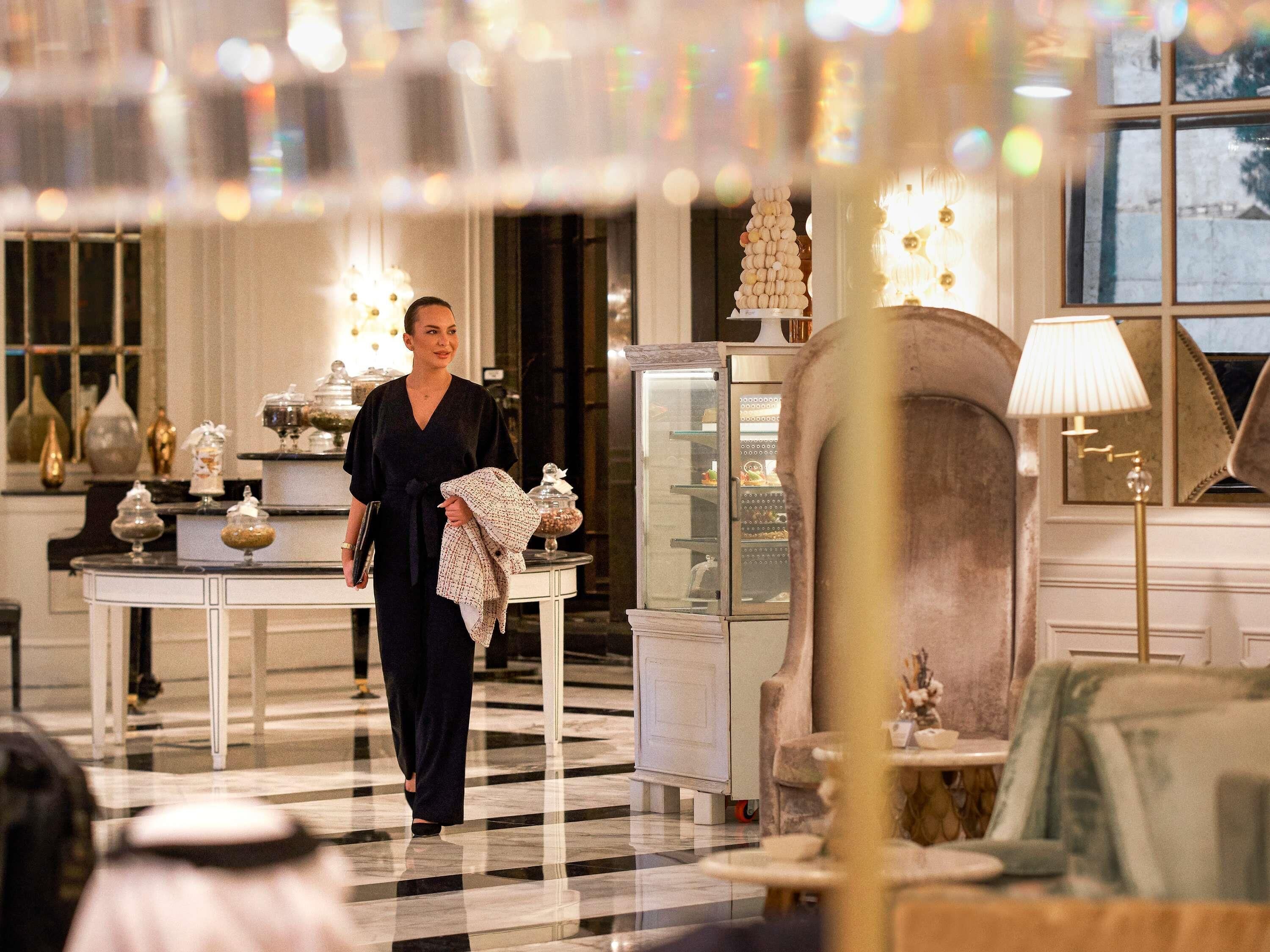 A woman walking through a hotel lobby with a checkered floor, a glass display case, and a lamp.