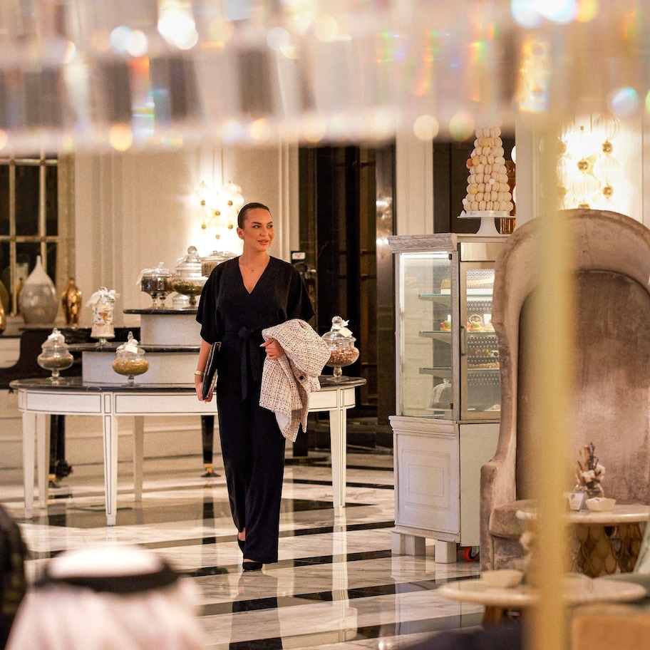 A woman walking through a hotel lobby with a checkered floor, a glass display case, and a lamp.