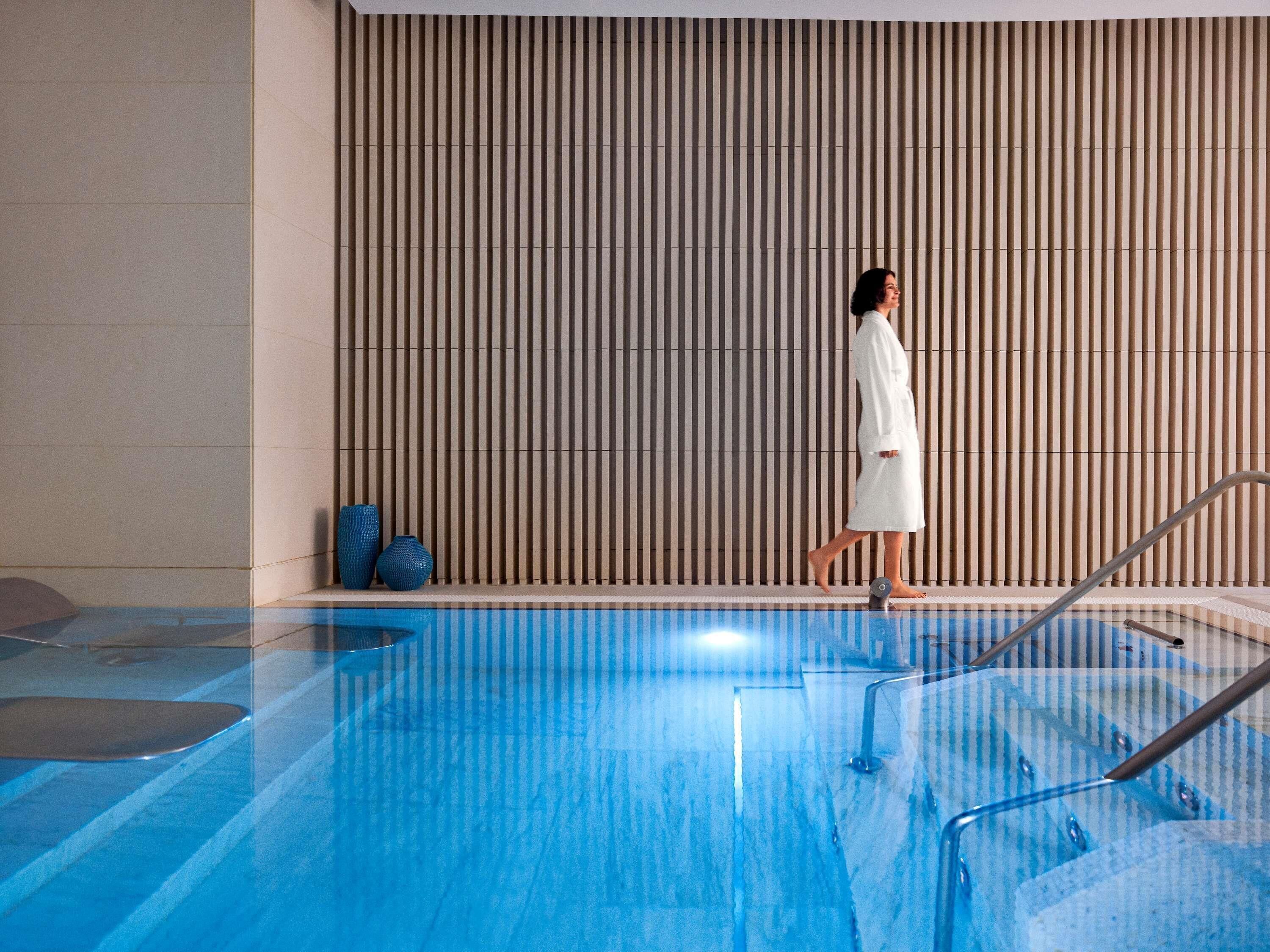 A person in a white robe walking by a pool with a wooden slatted wall in the background.