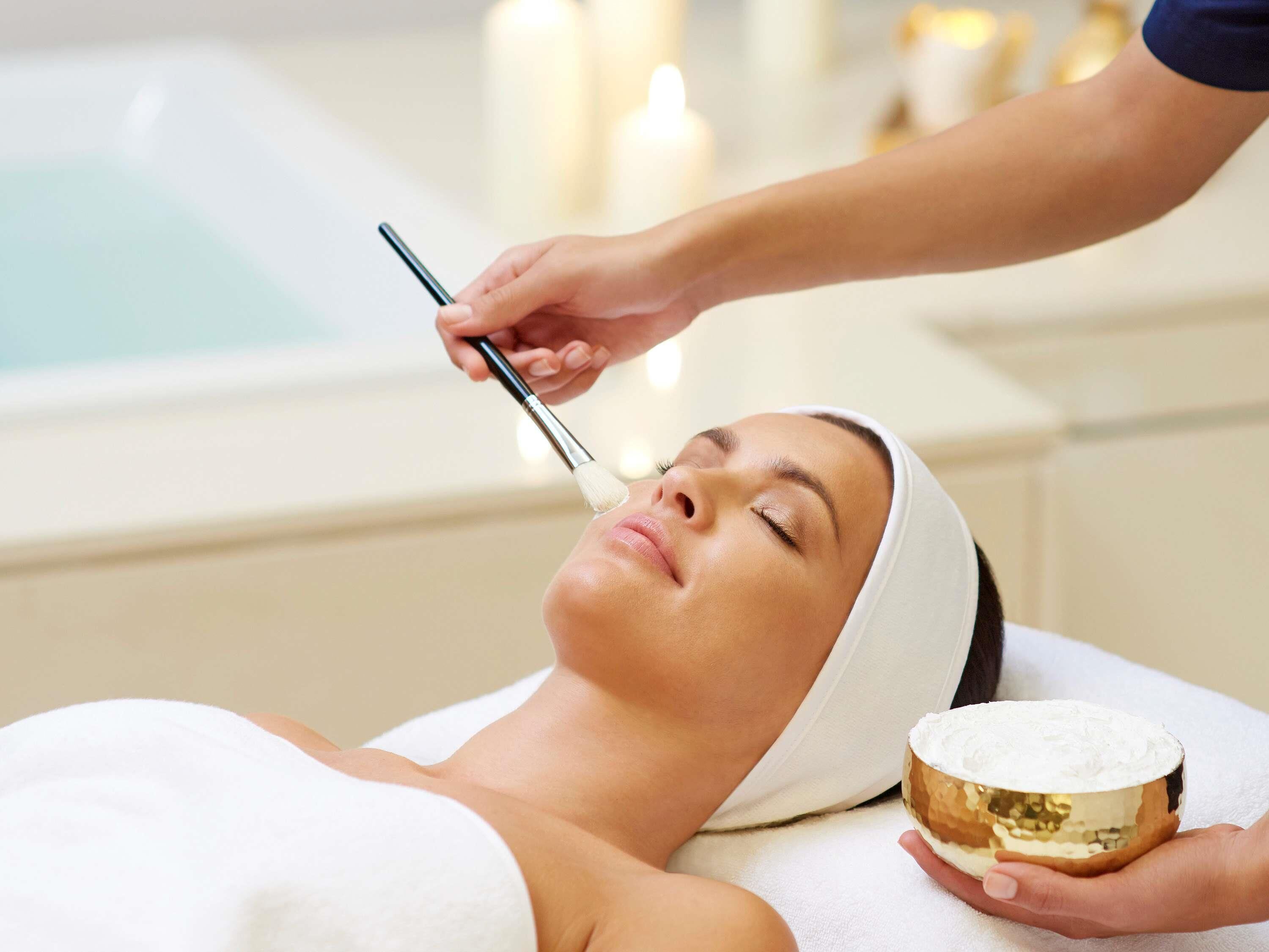 A person receiving a facial treatment in a spa setting with a bathtub and candles in the background.
