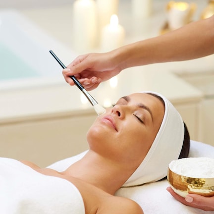 A person receiving a facial treatment in a spa setting with a bathtub and candles in the background.