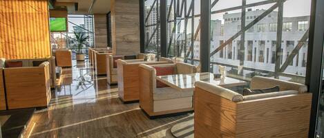 A modern hotel lobby with wooden furniture, large windows, and a flat-screen TV.
