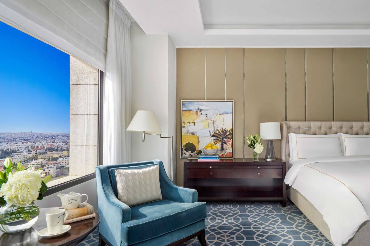 A hotel room with a large window, a blue armchair, a bed, a nightstand, and a painting on the wall.