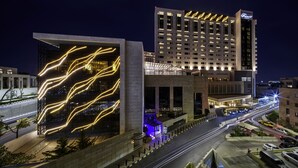 A modern hotel with a prominent facade featuring illuminated lines, a large parking area, and a busy street in the background. - Fairmont Amman (Amman)